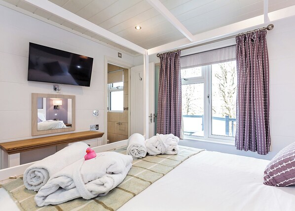 A hotel room with a bed, a TV mounted on the wall, a bathroom with a shower, and a window with curtains.
