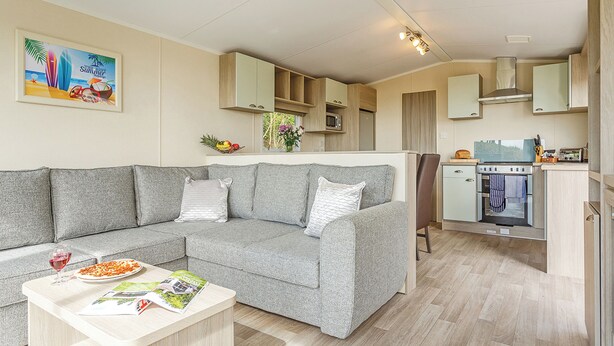 A modern living room with a grey sofa, a dining table with a magazine, and a kitchen area with wooden cabinets and a microwave. - 2 bedroom accommodation in Sandymouth Bay, Bude (Sandymouth Bay, Bude)