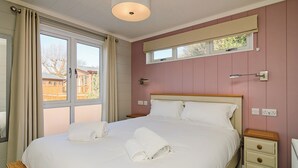 Un lit bien fait, avec du linge de lit et des serviettes blanches, une table de chevet, une commode, une fenêtre donnant sur un immeuble et une lampe fixée au mur.
