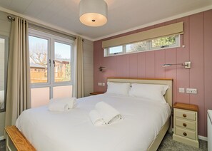 En snyggt bäddad säng med vita lakan och handdukar, ett sängbord, en liten avlastningsyta bredvid sängen, ett fönster med utsikt över en byggnad och en vägglampa.