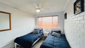 Een kamer met twee eenpersoonsbedden, een plafondventilator, een raam met jaloezieën en een witte bakstenen muur.