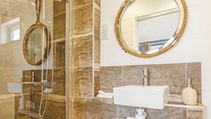 A bathroom with a glass-enclosed shower, a white sink, a round mirror with a rope frame, and a marble-tiled wall.