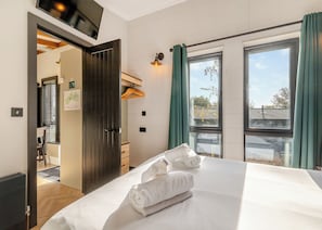 Un dormitorio con cama, ventana con cortinas, una puerta y una mesita.