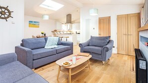 Un salon moderne avec un canapé, un fauteuil et une table basse, une cuisine en arrière-plan et une porte en bois.