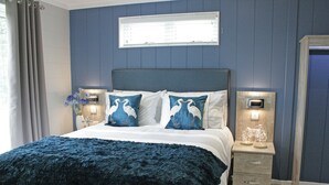 A bedroom with a bed, two bedside tables, a nightstand, a window with blinds, and a wall-mounted light fixture.