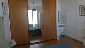 A room with a wooden wardrobe, a bed with a blue bedspread, a small white shelf, and a wall clock.