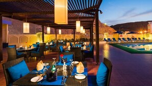 Un espace extérieur aménagé pour les repas, avec des tables pour deux, placé sous une pergola illuminée de guirlandes lumineuses, et un coin piscine avec des chaises longues en arrière-plan.