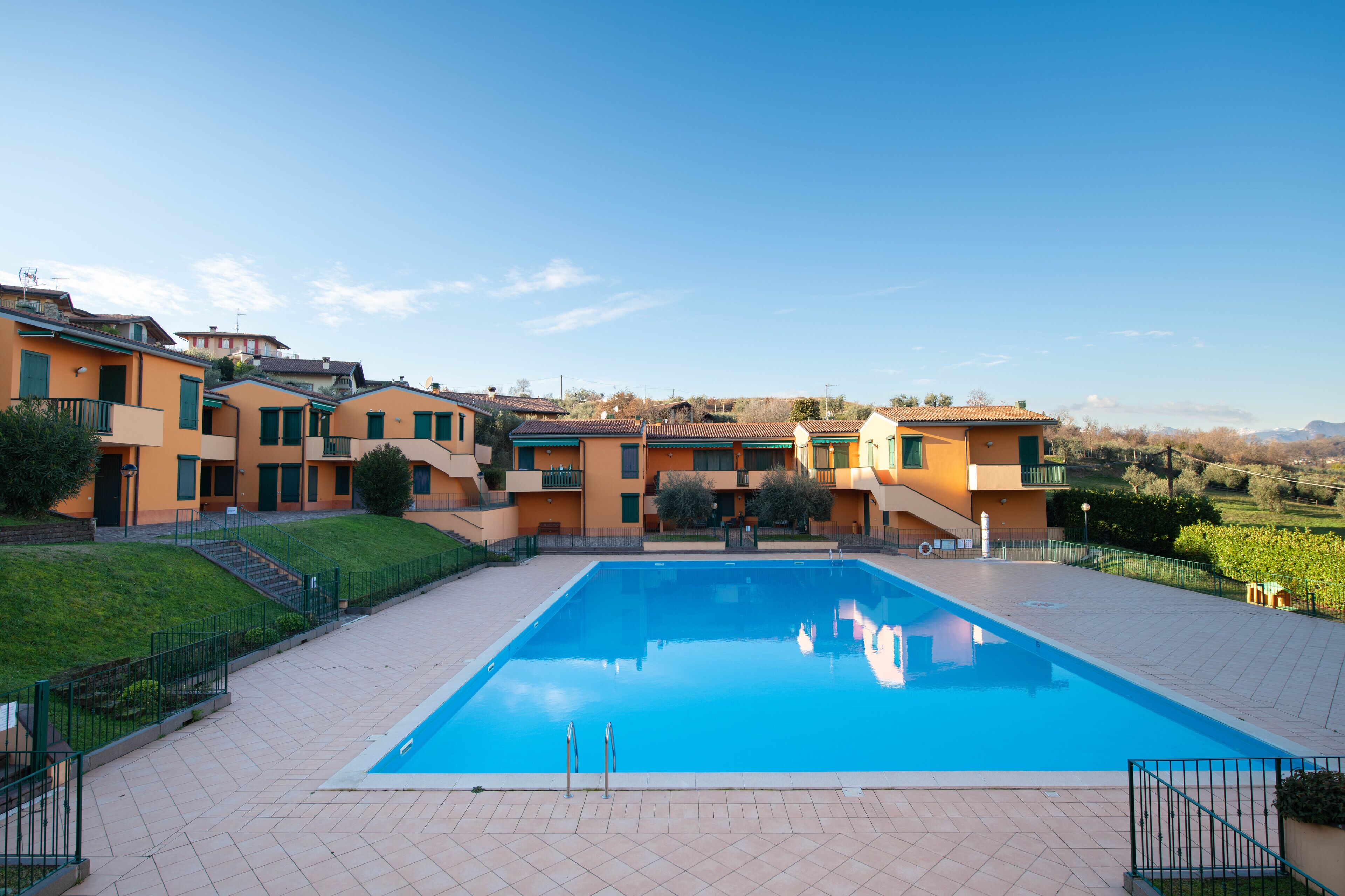 A swimming pool with a tiled deck, surrounded by multi-story buildings with balconies, set against a clear sky.