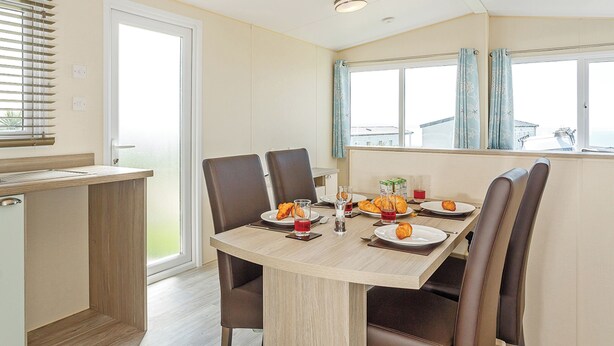 Un coin repas avec une table dressée pour quatre personnes, des chaises, une fenêtre avec des rideaux et un petit coin cuisine.
