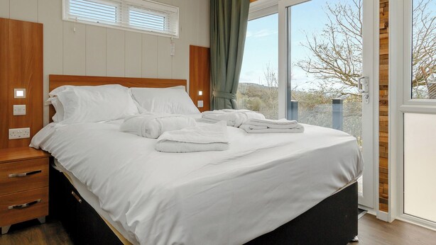 A hotel room with a bed, bedside tables, a window with curtains, and a view of the outdoors. - This charming one-bedroom lodge boasts all the home comforts you need. (Cheddar)