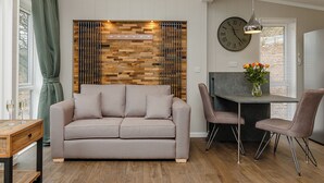 A modern living room with a sofa, dining table, chairs, a clock, and a decorative wall panel. - This charming one-bedroom lodge boasts all the home comforts you need. (Cheddar)