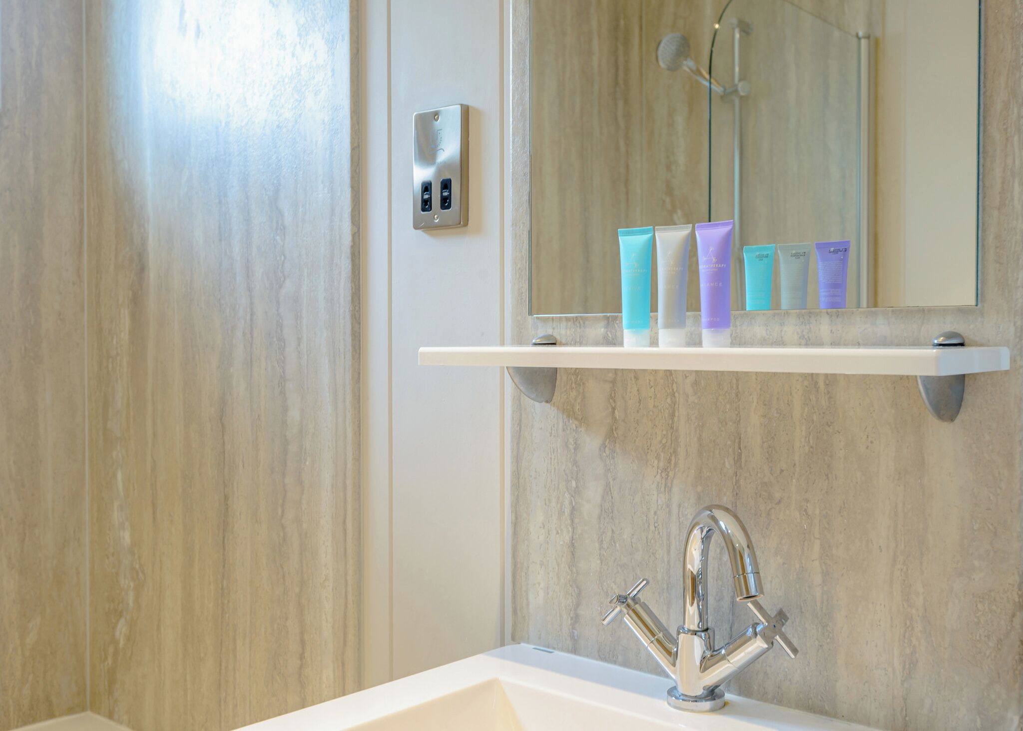 Un baño con lavamanos blanco, grifo cromado, espejo y repisa con artículos de aseo.