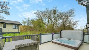 Una terraza de madera con un jacuzzi, mobiliario de mimbre y vistas a una casa y árboles.