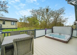 Una terraza de madera con un jacuzzi, mobiliario de mimbre y vistas a una casa y árboles.