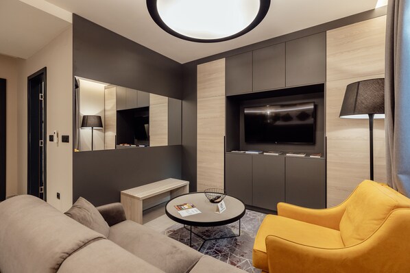 Quarto de hotel moderno com sofá, poltrona amarela, mesa de centro redonda, televisão e um banco.