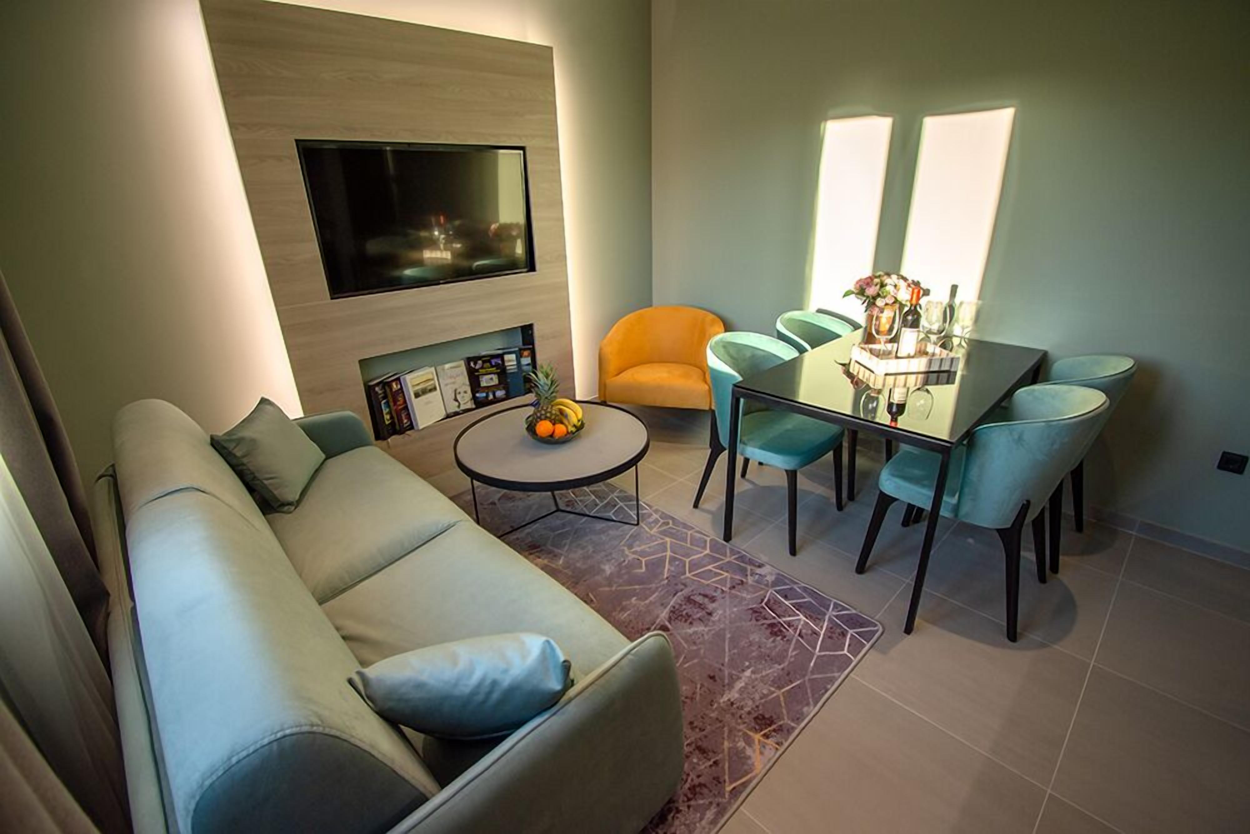 A modern living room with a sofa, a dining table with chairs, a flat-screen TV mounted on the wall, and a small round table with fruits.