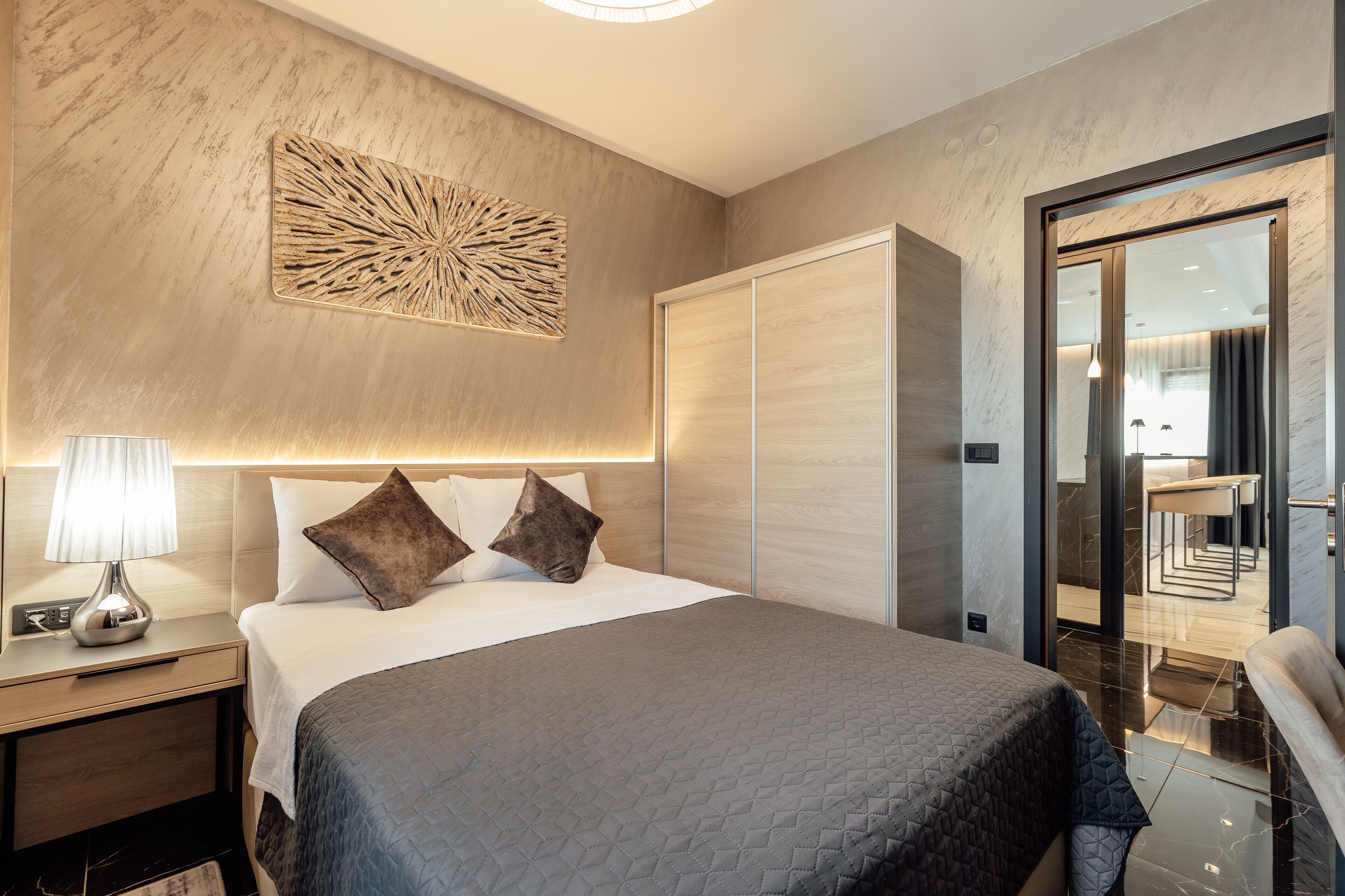 Een moderne hotelkamer met een groot bed, een nachtkastje met een lamp, een decoratief kunstwerk en zicht op het interieur van het hotel door een glazen deur.