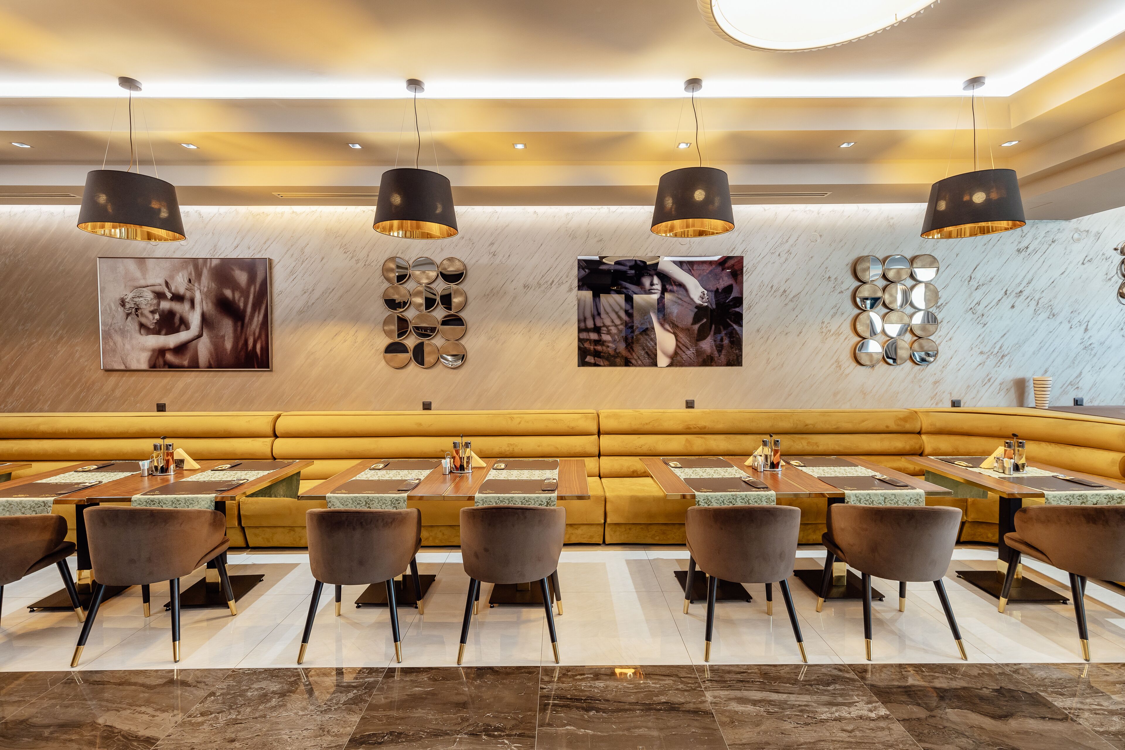 Een modern restaurant met een lange, gele zithoekbank, verschillende tafels met stoelen, en decoratieve muurkunst.