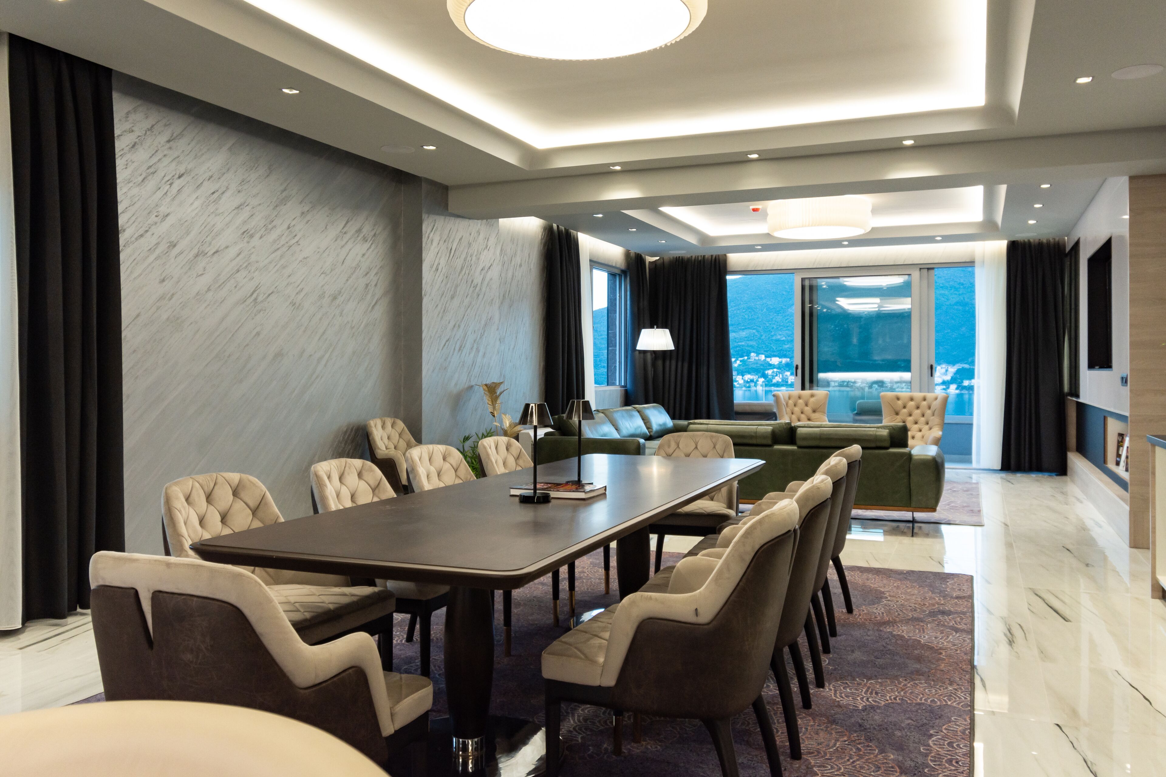 A modern hotel room with a dining area, a sofa set, and a large window offering a city view.