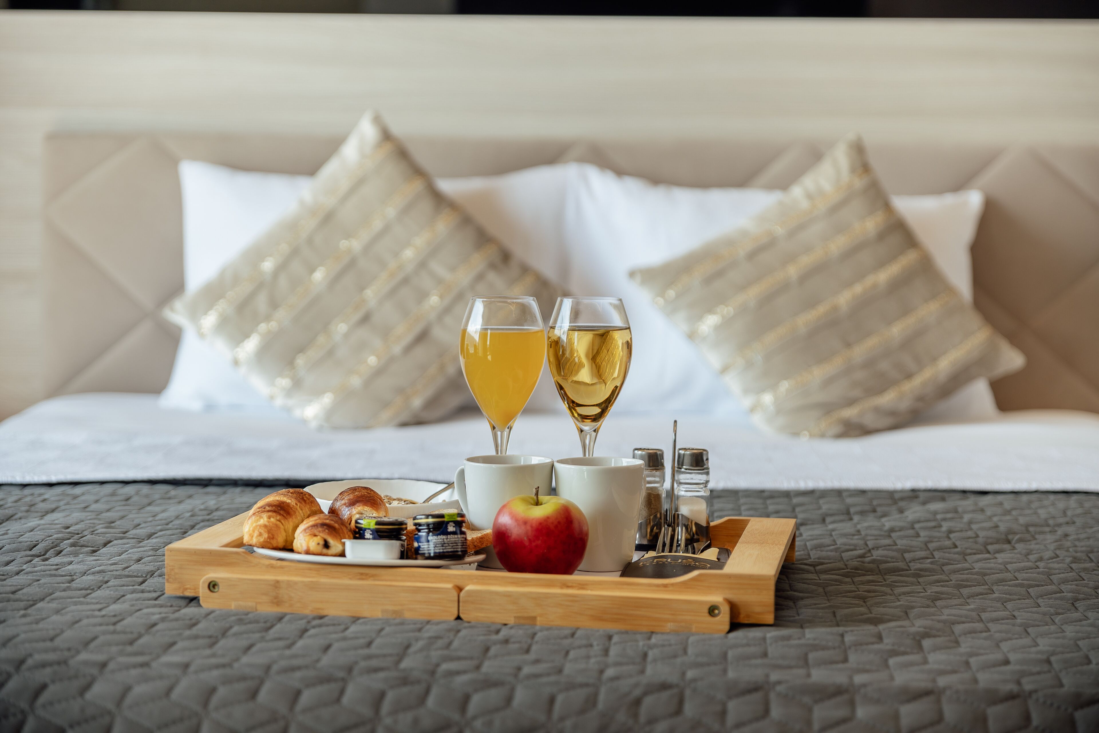 Een ontbijfwagen met croissants, een appel en twee glazen sinaasappelsap op een bed met een gewatteerde hoes en decoratieve kussens.