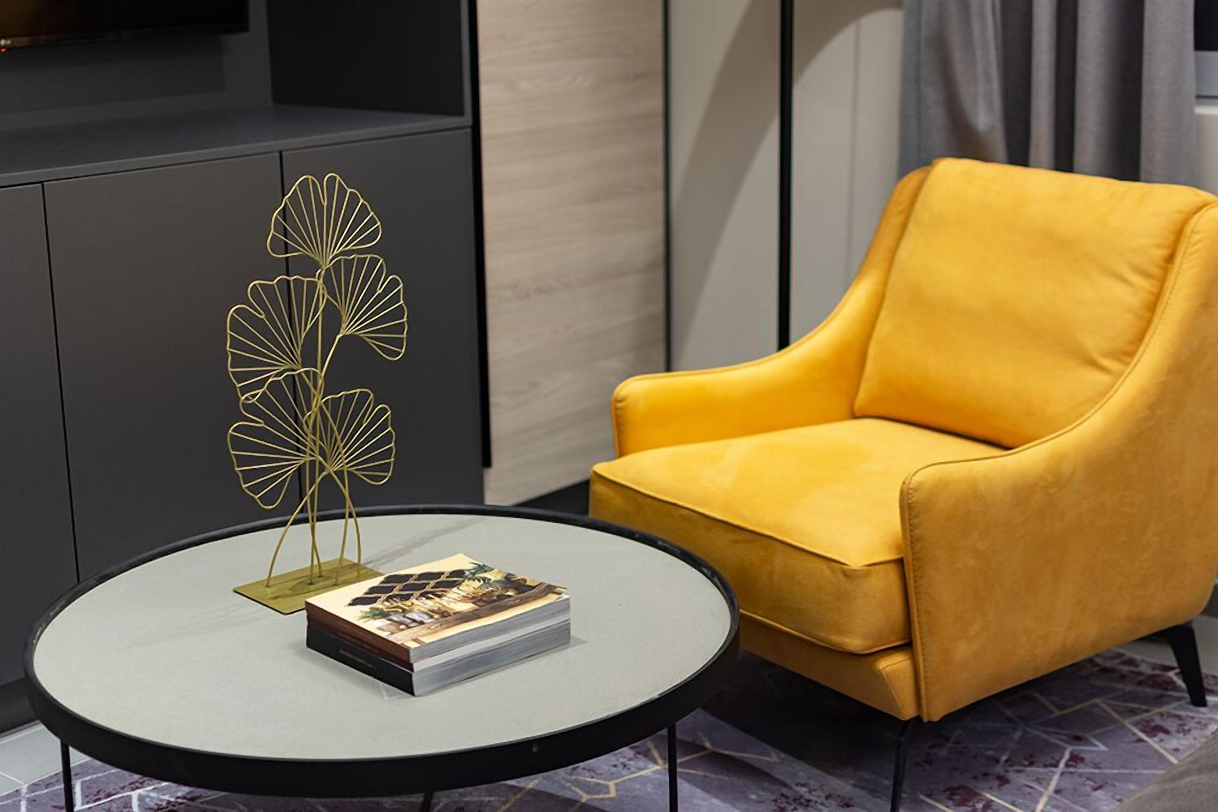 Un fauteuil jaune placé à côté d’une table ronde, sur laquelle se trouvent une plante décorative en métal et un livre.