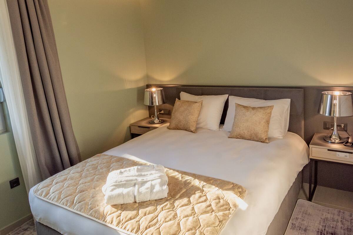 A well-lit hotel room with a large bed, two bedside tables with lamps, a ceiling light, and a window with curtains.