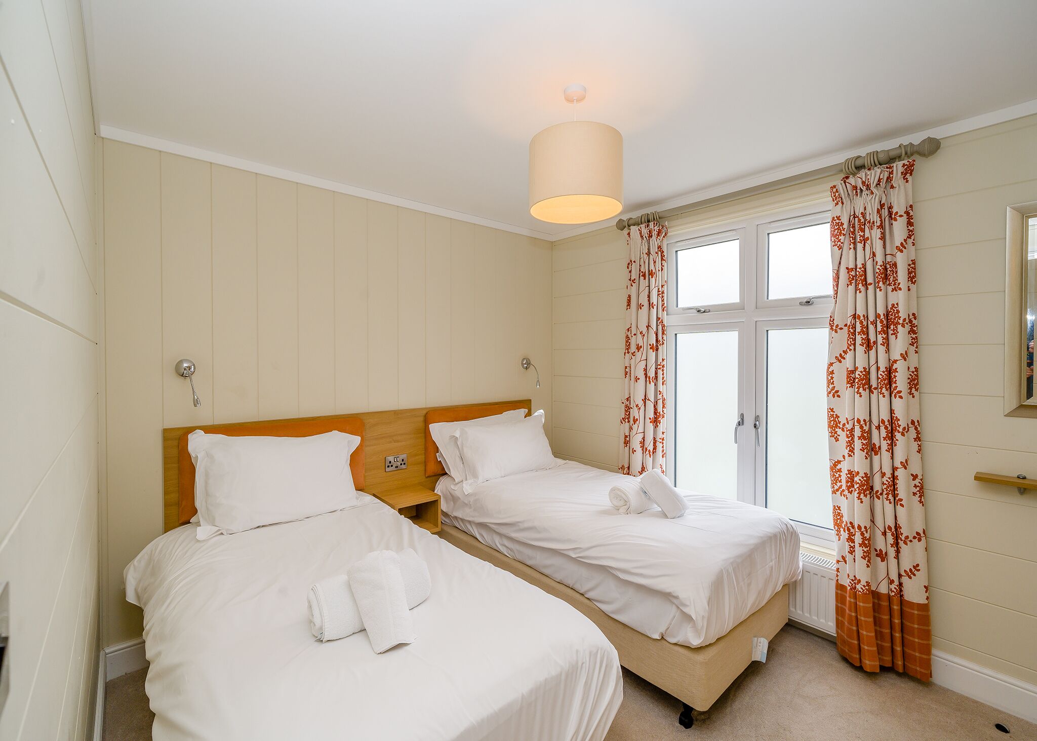 A hotel room with two beds, a wooden headboard, a ceiling light, a window with curtains, and a radiator.
