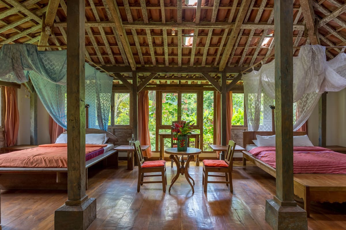 Ein Zimmer mit Holzbalken, einem Bett mit roter Decke, einem Tisch mit Stühlen und einem Fenster mit Vorhängen.