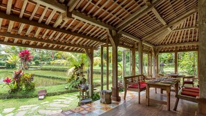 A covered outdoor area with wooden flooring, tables, and chairs, surrounded by a lush garden and a pond.