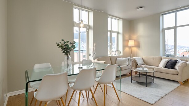 A modern living room with a glass dining table, white chairs, a beige sofa, and a view of the city through large windows.