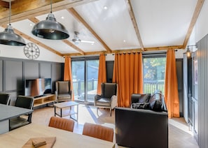 A modern living room with a dining area, large windows with orange curtains, a ceiling fan, and a clock on the wall.