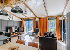 A modern living room with a dining area, large windows with orange curtains, a ceiling fan, and a clock on the wall. - Elkstone Premier Lodge (Ashbourne)