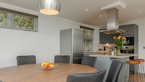 A modern dining area with a round wooden table, grey upholstered chairs, a fruit bowl, and a kitchen in the background. - 4 bedroom accommodation in Cheddar (Cheddar)