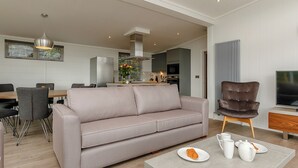 A modern living room with a sofa, a coffee table with a croissant, and a TV. - 4 bedroom accommodation in Cheddar (Cheddar)