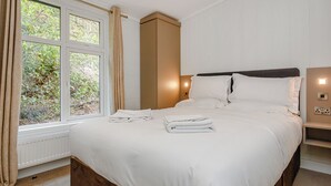 A bedroom with a large bed, a bedside table, a nightstand, a window with curtains, and a view of greenery outside. - 4 bedroom accommodation in Cheddar (Cheddar)