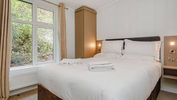 Ein Schlafzimmer mit einem großen Bett, einem Nachttisch, Vorhängen am Fenster und Blick ins Grüne.