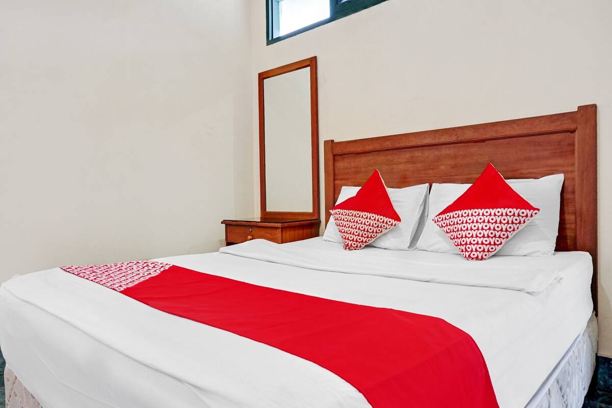 A neatly made bed with a red and white bedspread, two pillows with a red pattern, a wooden headboard, and a nightstand with a mirror.