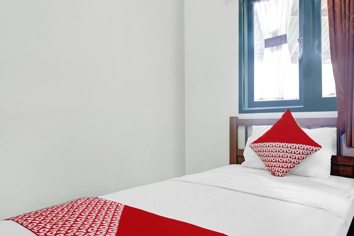 A single bed with a white and red bedspread, a wooden headboard, and a window with curtains.