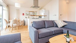Un salon moderne avec un canapé gris, une table basse en bois et un coin cuisine à l’arrière-plan.