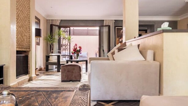 Un salon moderne avec un canapé, un pouf et une table basse. Il y a un téléviseur sur la gauche et une fenêtre avec des rideaux.