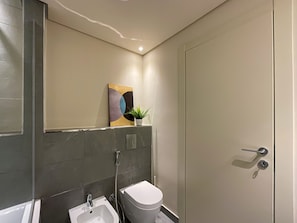A modern bathroom with a toilet, bidet, and sink. There is a potted plant on the wall and a painting above the sink. - WelHome - Evergreen Apartment Amidst Vibrant JVC With Pool (Dubai)