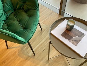 A green upholstered chair, a round table with a book and a candle, and a wooden floor. - WelHome - Evergreen Apartment Amidst Vibrant JVC With Pool (Dubai)