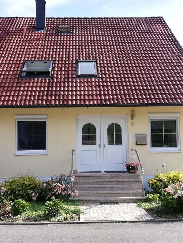 A building with a red-tiled roof, a double door entrance, and a small garden with flowers.