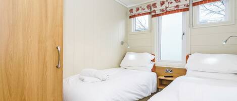 A room with two beds, a wooden wardrobe, a window with curtains, and a bedside table with a lamp.