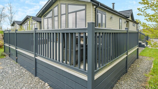 A modern caravan with a large windowed front section, a dark grey metal fence, and a gravel driveway. - 2 bedroom accommodation in Bewaldeth, Keswick (Bewaldeth, Keswick)