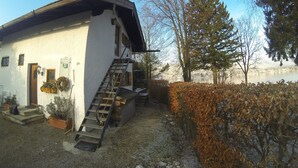 A white building with a wooden door, a staircase leading to a second floor, and a view of a lake with trees in the background. - Small apartment for a maximum of 2 people in a unique location directly on the lake (Rottach-Egern)