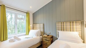 A bedroom with two beds, a wooden nightstand, a window with curtains, and a headboard with a plaid pattern. - 1 bedroom accommodation in Holton Heath (Holton Heath)
