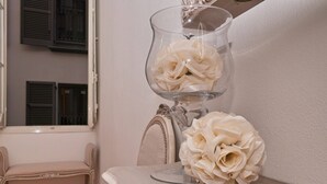 Une pièce avec un vase en verre contenant des fleurs blanches, posé sur une table, une chaise et une fenêtre avec des volets.