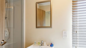 Ein Badezimmer mit Duschkabine aus Glas, einem weißen Waschbecken mit Toilettenartikeln, einem Spiegel und einem Fenster mit Jalousien.