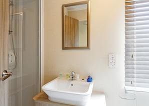 A bathroom with a glass shower enclosure, a white sink with toiletries, a mirror, and a window with blinds.